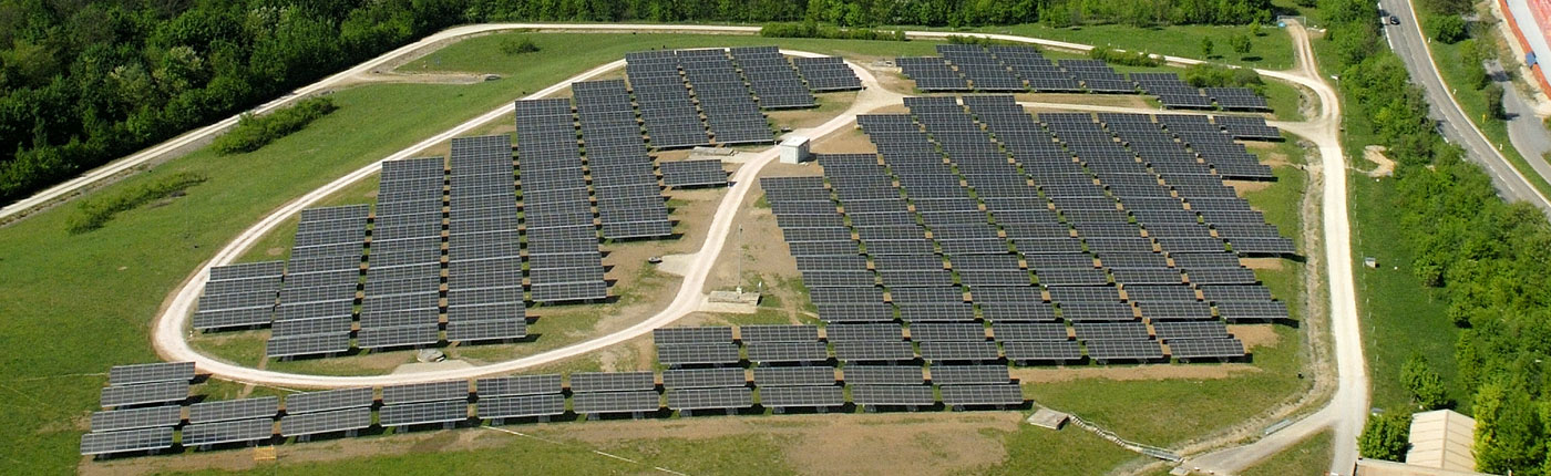 Luftaufnahme bei Tag der Deponie in Malsch im heutigen Zustand. Zu sehen ist die Phtovoltaikanlage. Sie steht auf einer Lichtung, um diese ein asphaltierter Weg herum führt. Die Photovoltaikanlage ist in zwei Felder aufgeteilt. Das linke Feld hat einen zusätzlichen Weg, der um diesen Bereich im Kreis herum führt. Rechts von der Anlage ist ein Teilstück einer Landstrasse zu sehen. Auf der linken Seite des Feldes ist ein Waldstück zu erkennen. In der Mitte der Photovoltaikanlage ist ein kleines Häuschen zu sehen, welches die Anschlusstechnik beinhaltet. Eine Photographie die, die Deponie in Malsch aus der Luft zeigt